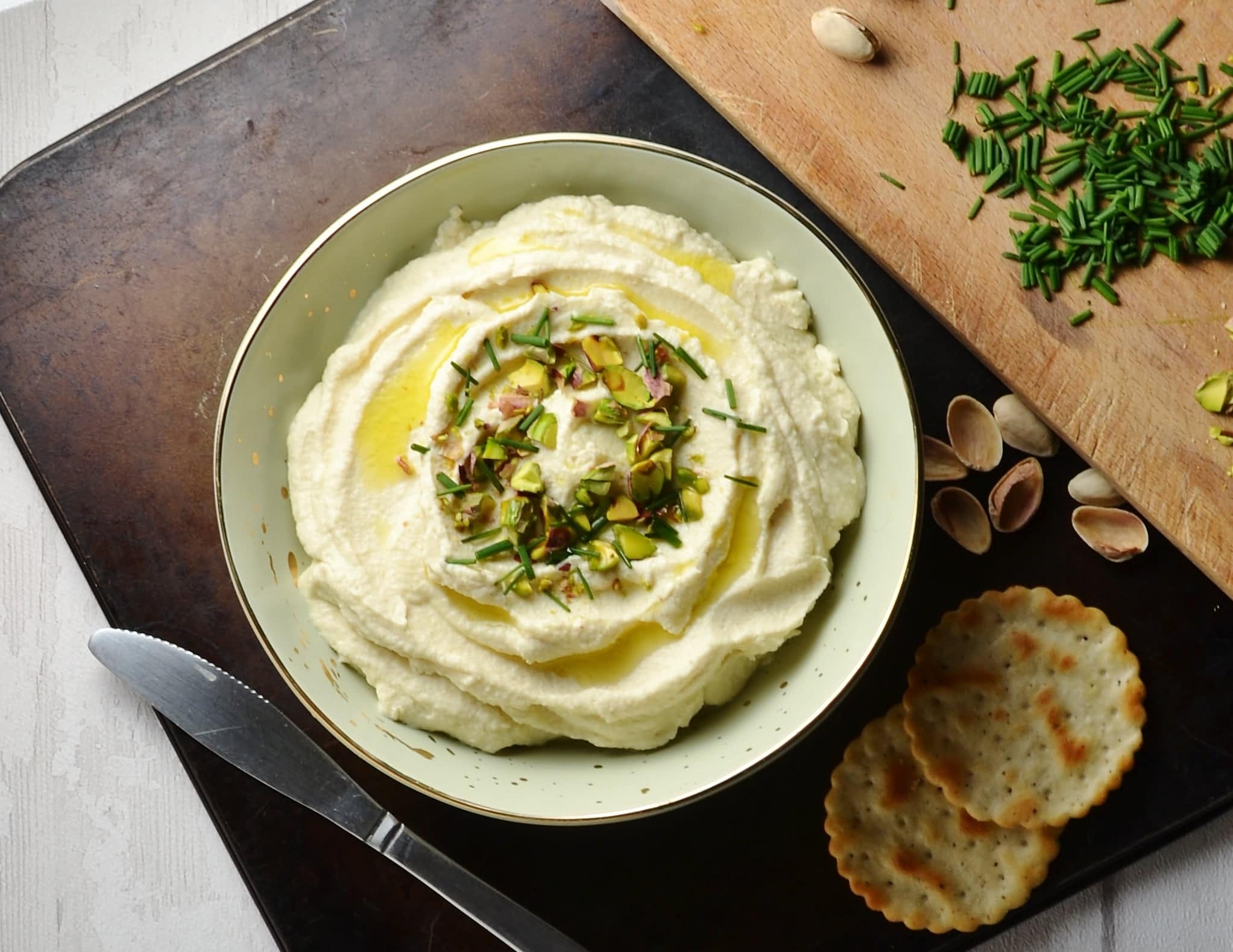 Top down view of cauliflower dip in creamy bowl on top of metallic tray with herbs and crackers in background.