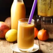 Sweet potato smoothie in glass with purple straw, lemon, tangerine, mango, smoothie in bottle and blender in background.