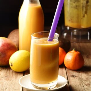Sweet potato smoothie in glass with purple straw, lemon, tangerine, mango, smoothie in bottle and blender in background.