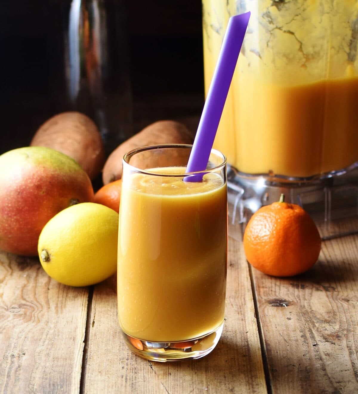 Sweet potato yellow smoothie in glass with purple straw, with tangerine, lemon, mango, sweet potatoes and smoothie in blender in background.