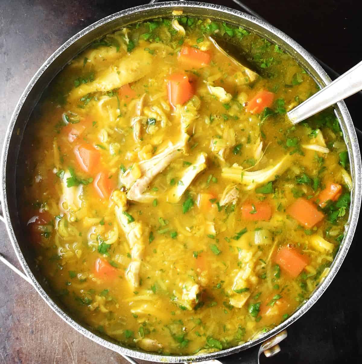 Top down view of chunky curried soup with chicken in large pot with ladle.