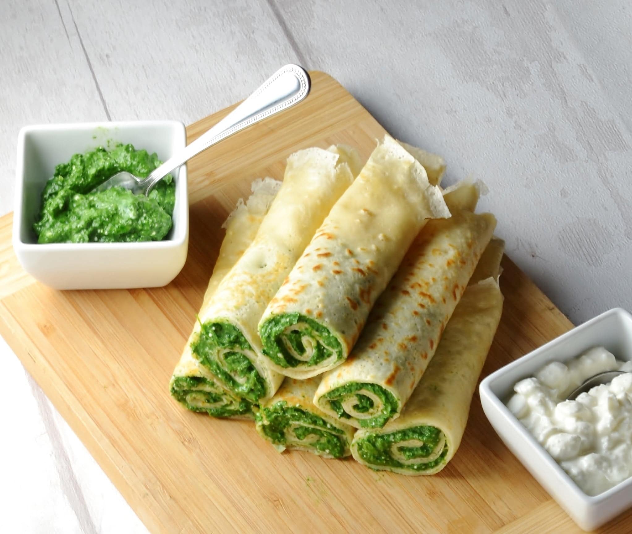 Crepes with spinach on wooden board with cottage cheese in white dish in bottom right corner and spinach mixture with spoon in white dish to left.