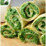 Crepes with spinach filling on wooden board with spinach mixture with spoon in white dish to left.