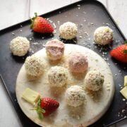 Strawberry cheesecake balls on round marble plate with strawberries and white chocolate on oven tray.