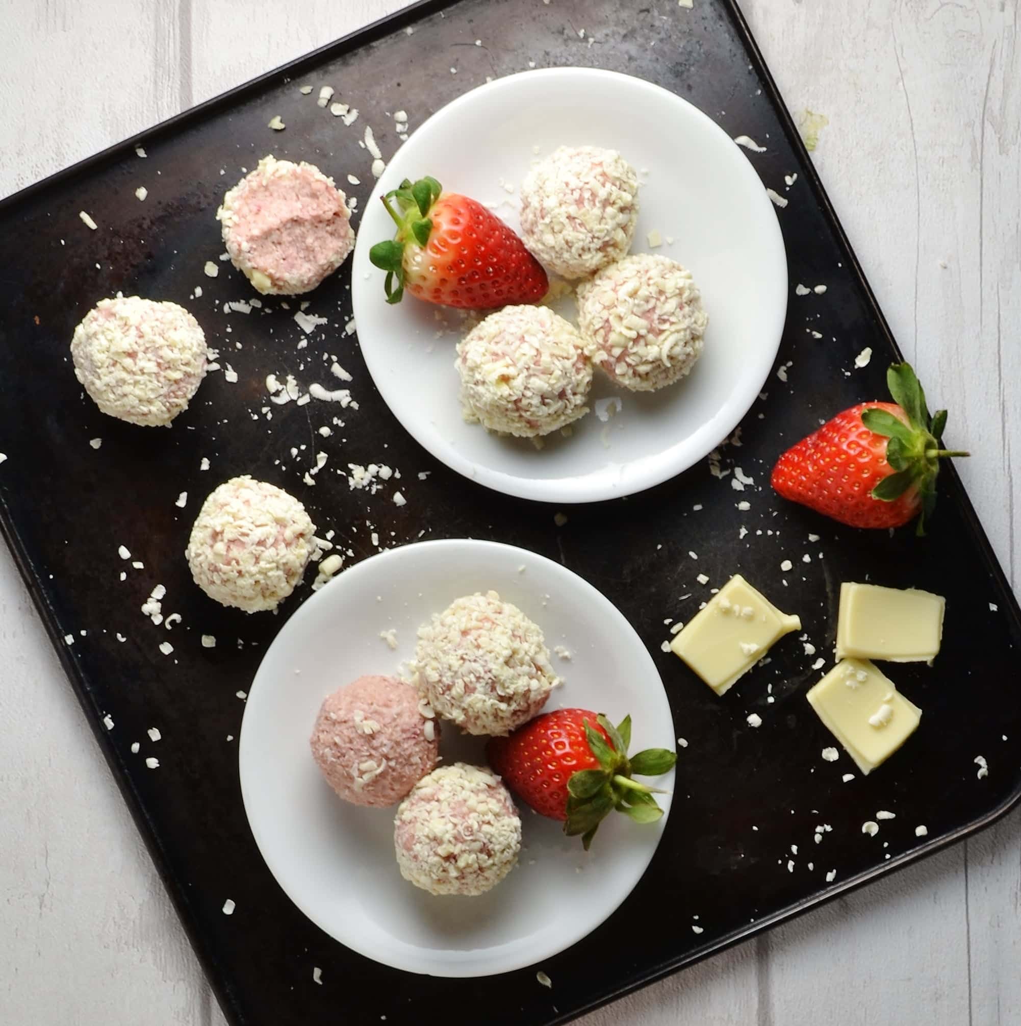Healthy strawberry cheesecake bites on two small white plates with white chocolate and strawberries on dark oven tray.