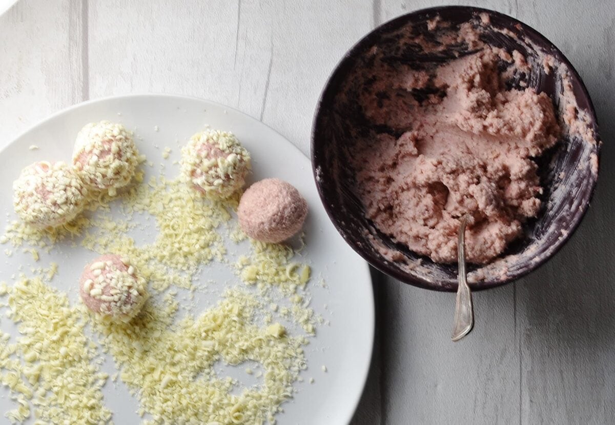 Strawberry cheesecake balls on white plate with white chocolate shavings and purple bowl with cheese mixture and spoon.