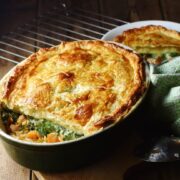 Salmon, spinach and sweet potato pie with puff pastry topping in green oval dish, with green cloth, spoon and pie in white bowl in background.