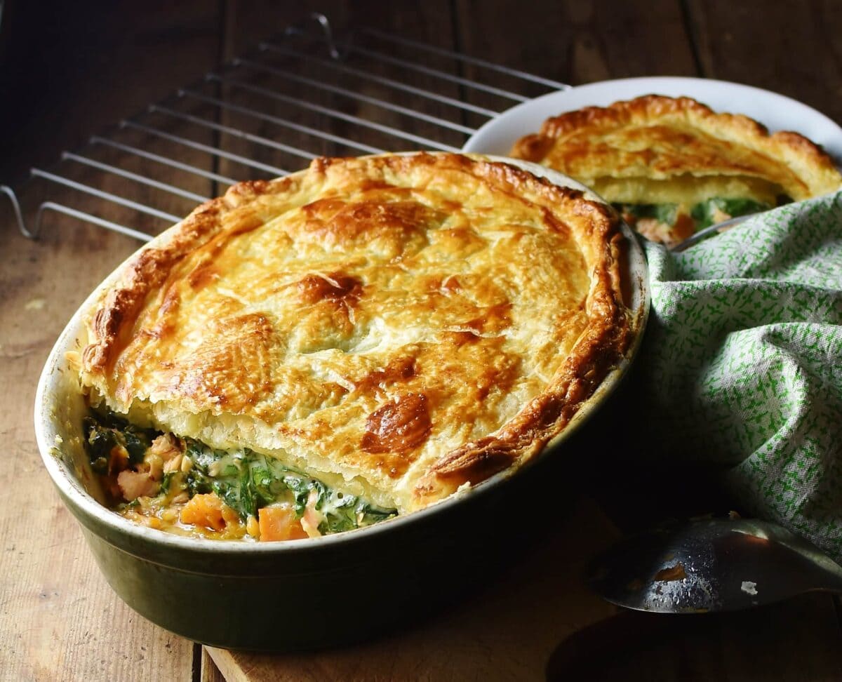 Salmon, spinach and sweet potato pie with puff pastry topping in green oval dish, with green cloth, spoon and pie in white bowl in background.
