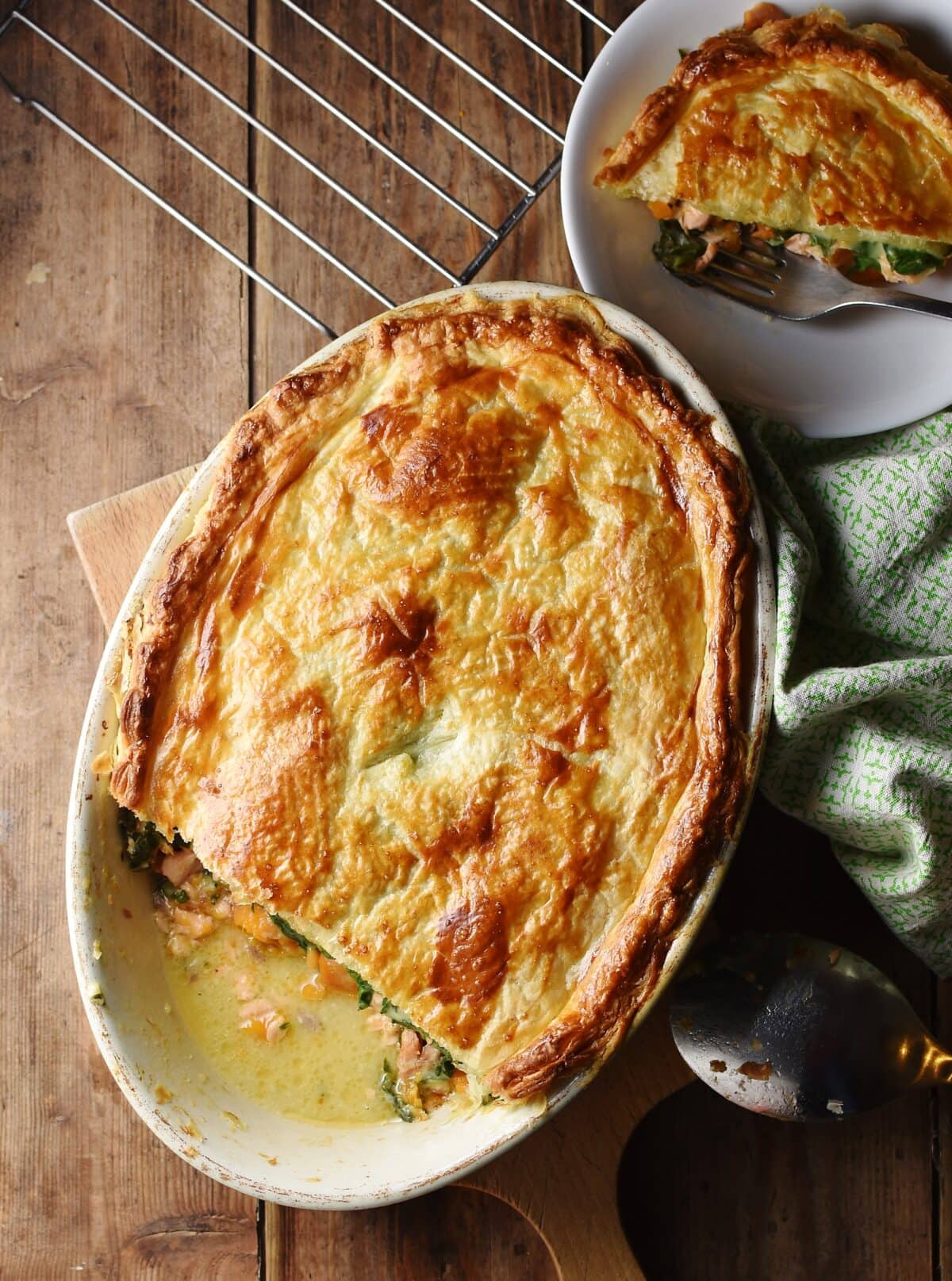Salmon pie with golden brown pastry in white oval dish, green cloth to the right and pie in white bowl in top right corner.