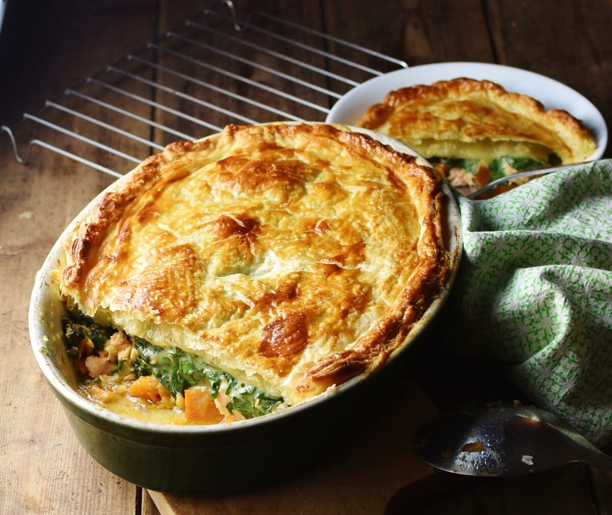 Side view of salmon spinach pie with golden puff pastry on top in oval dish, with green cloth to the right and pie portion in white bowl in background.