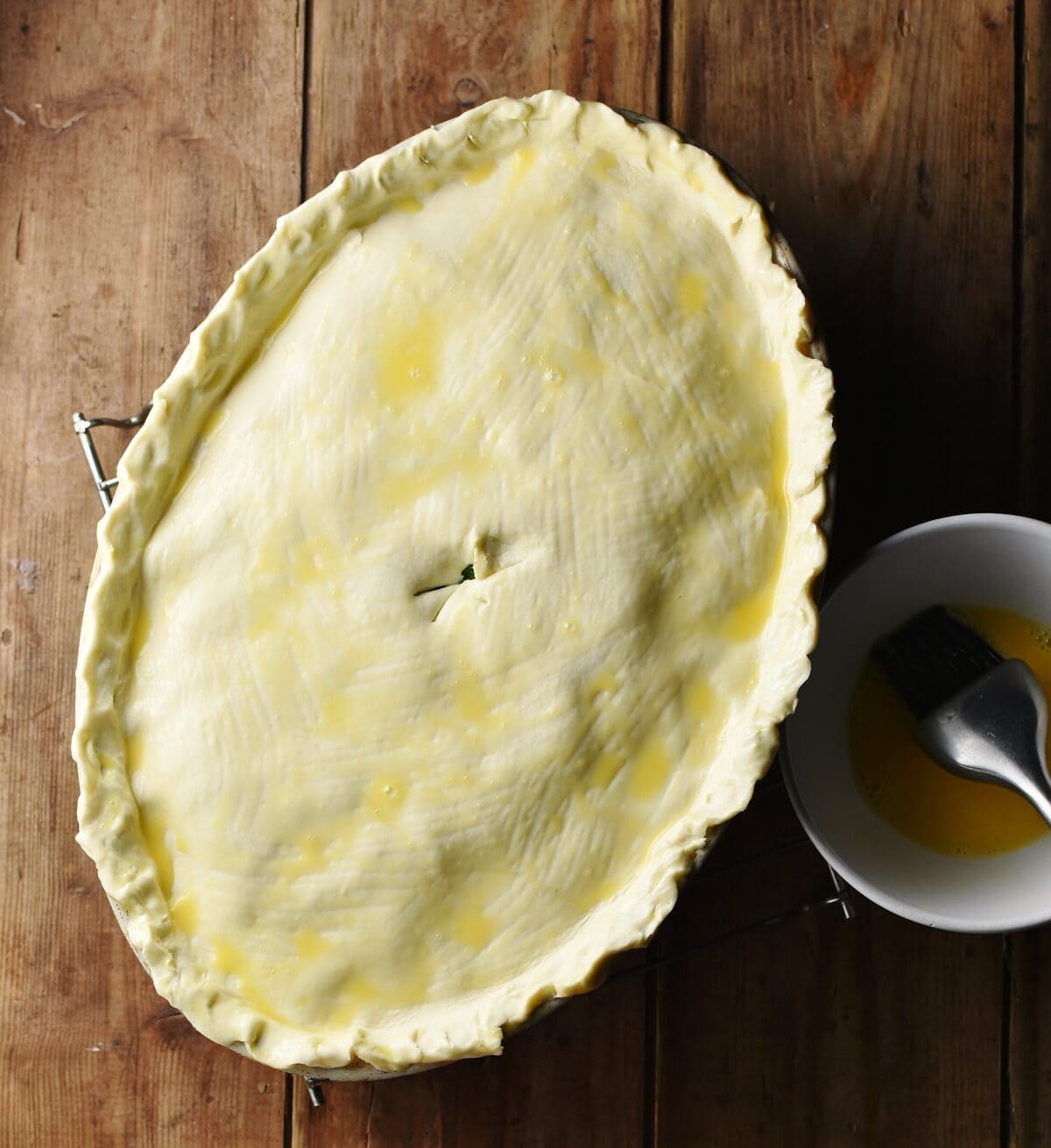 Pie covered with raw pastry in oval dish, with egg wash and brush to the right.