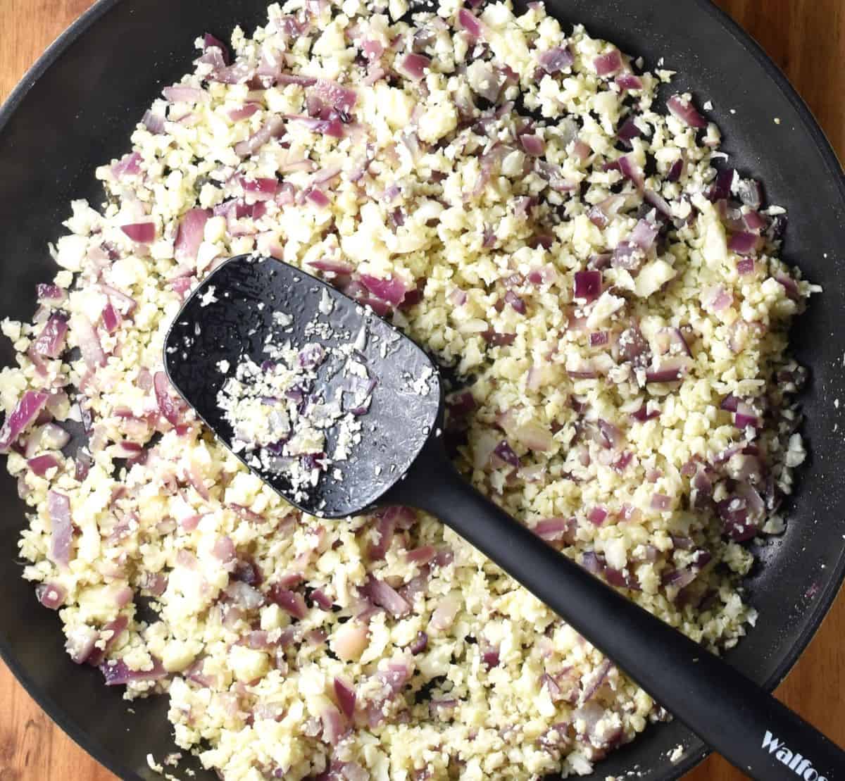 Cauliflower rice and chopped onion in large pan with black spatula.