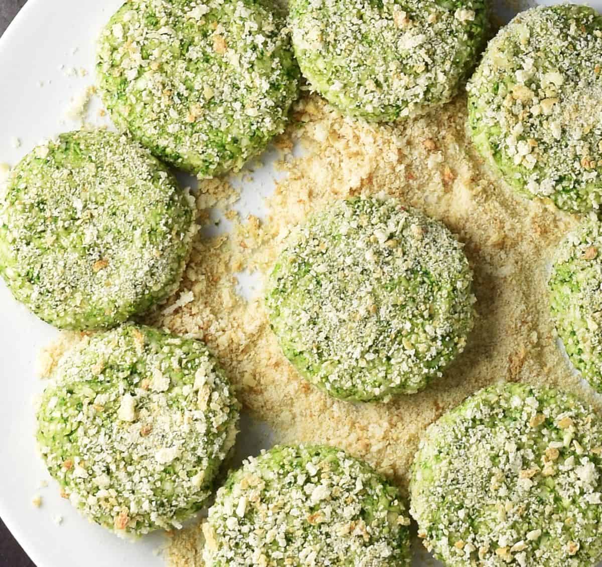 Uncooked breaded broccoli patties on white plate with breadcrumbs.
