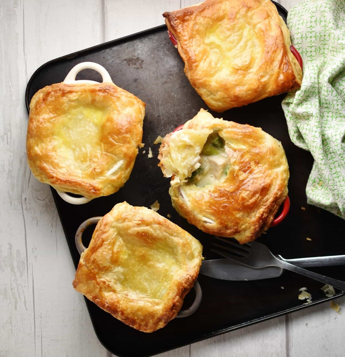 Top down view of mini chicken pot pies with knife, fork and green cloth to right on top of dark tray.