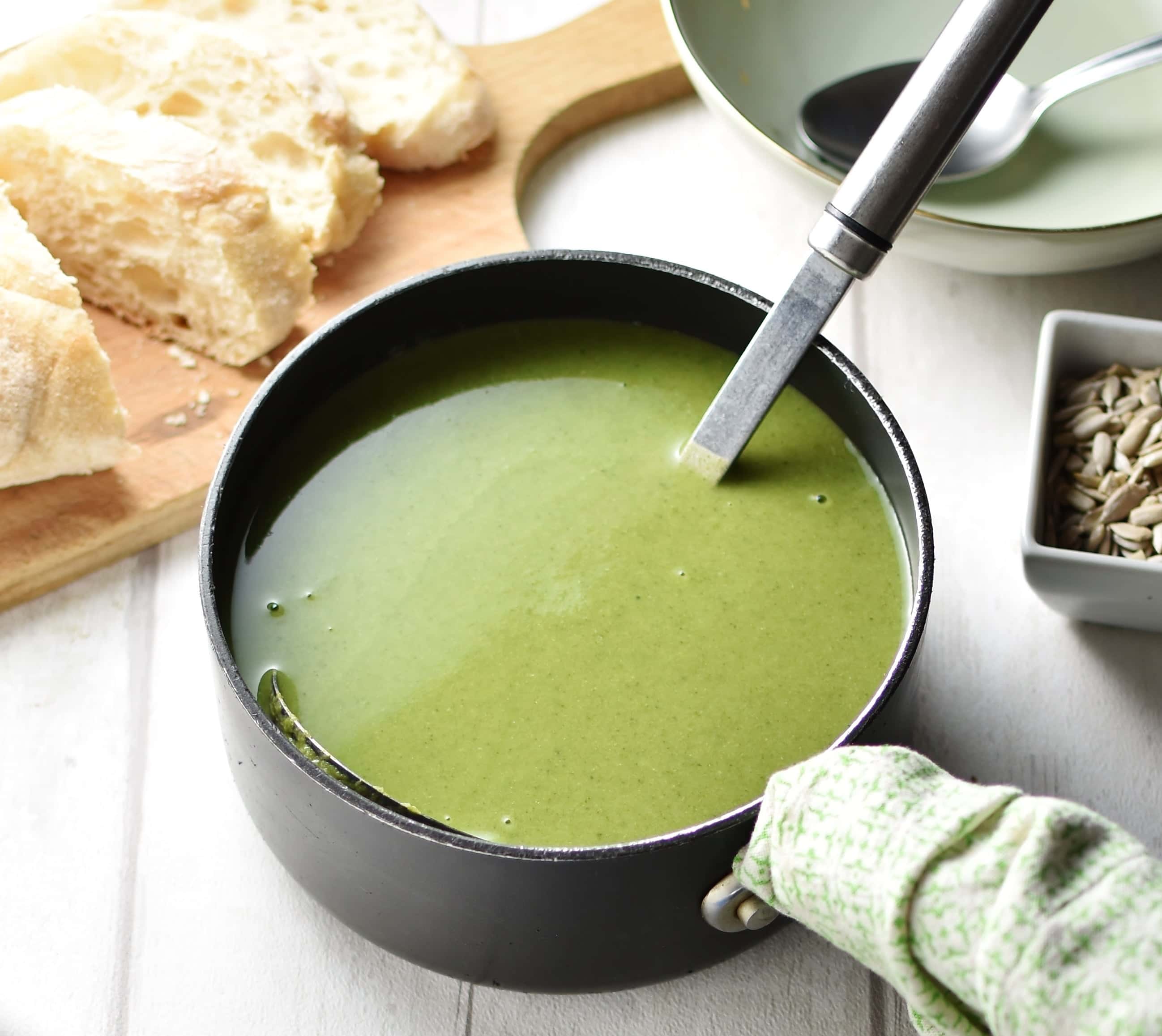 Broccoli spinach soup with ladle in black pot with green cloth wrapped round handle, bowl with spoon and slices of bread on top of wooden board in background.