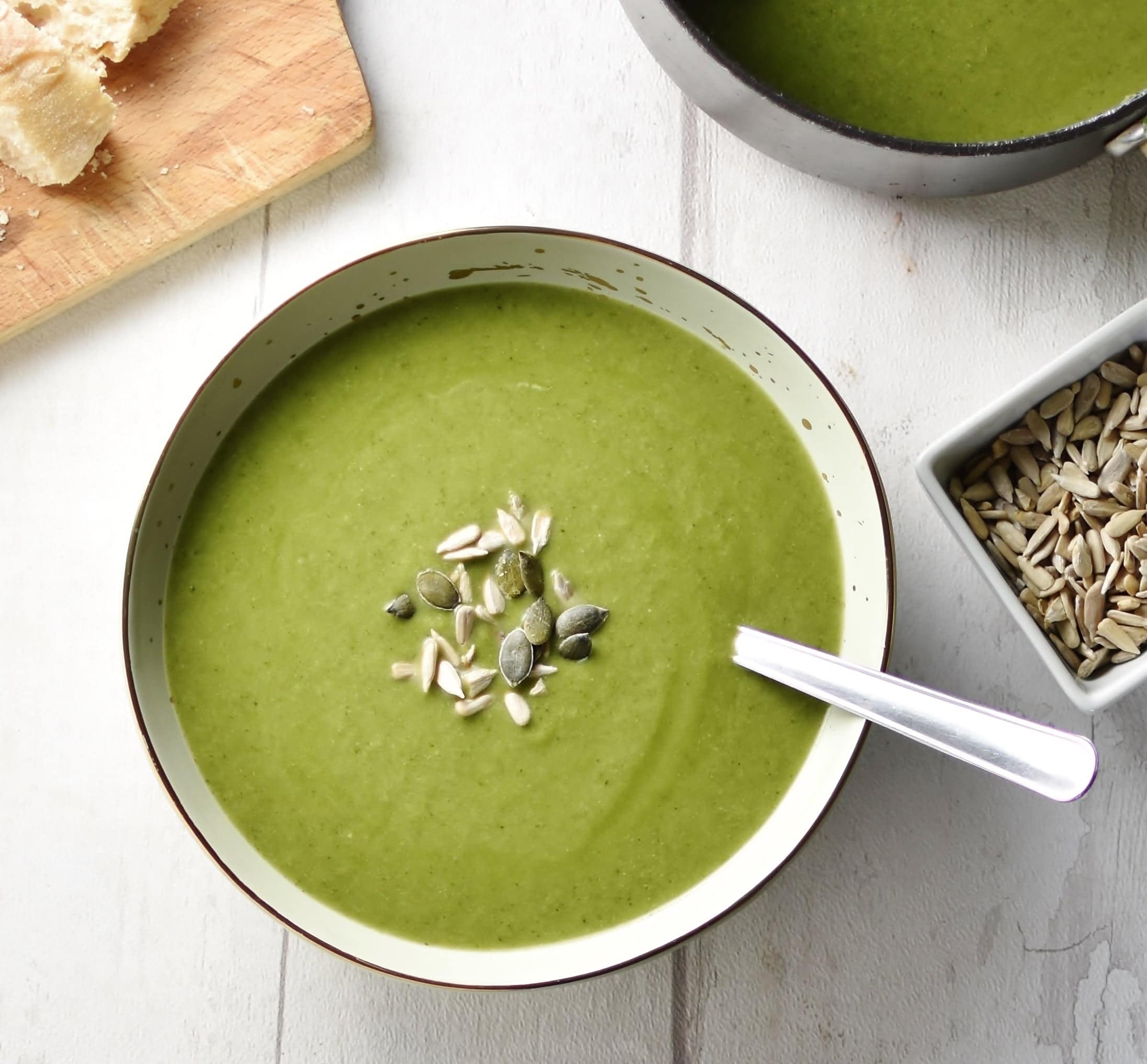 Top down view of broccoli spinach soup with seeds in grey bowl with spoon, seeds and saucepan with soup to right and wooden board in top left corner.