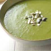 Partial side view of creamy broccoli soup with pumpkin seeds in green bowl on top of white board.