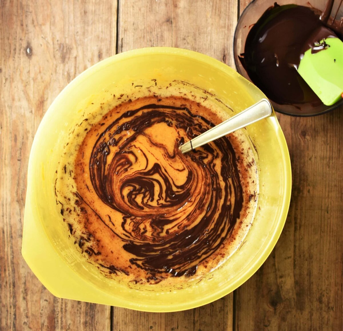 Sweet potato brownie mixture with melted chocolate in large yellow bowl with spoon and melted chocolate with green spatula in small dish in top right corner.