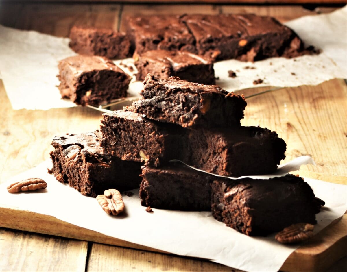Brownies stacked on top of paper with pecans and brownies in background.