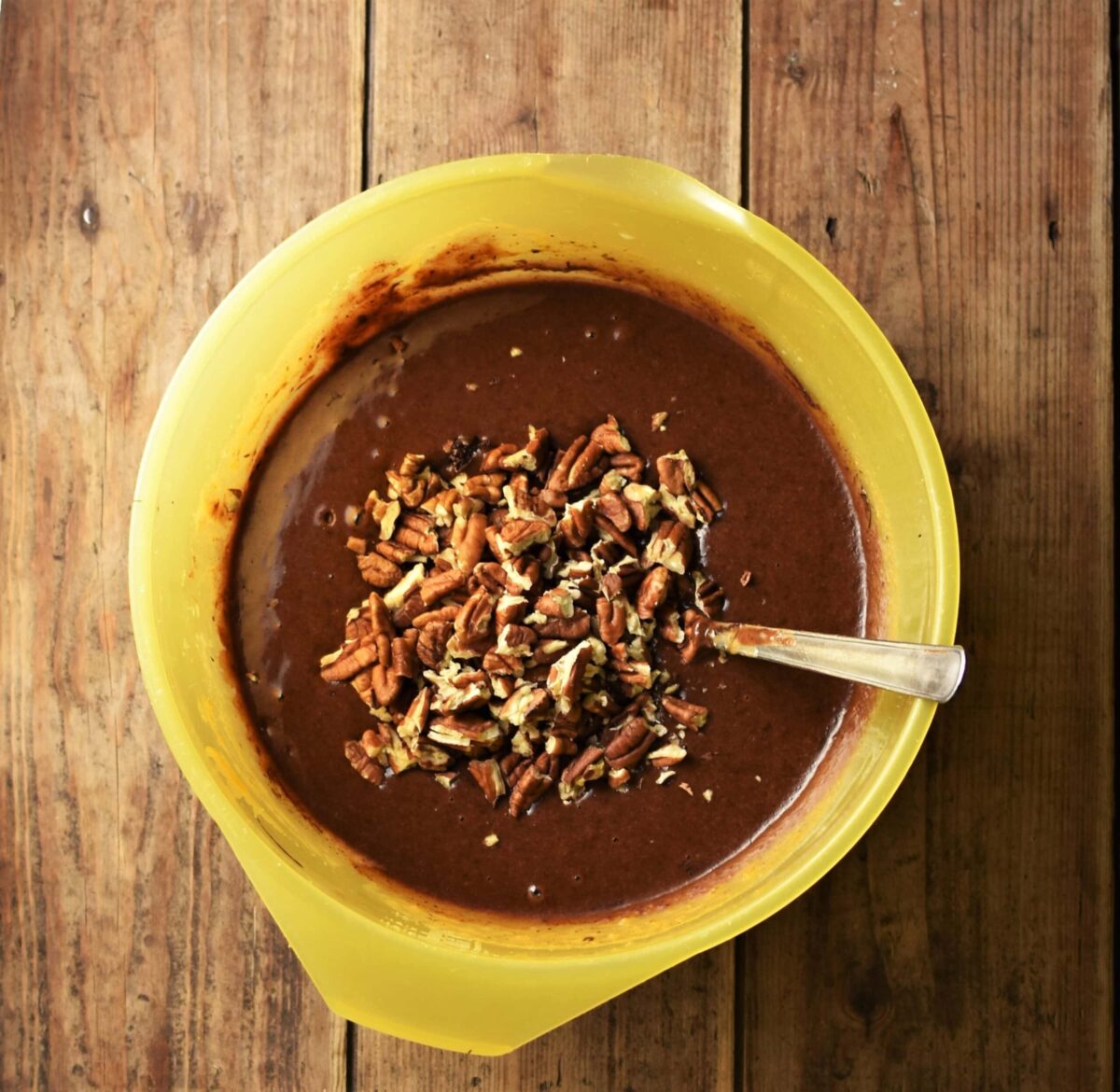 Brownies batter with chopped pecans in large yellow bowl with spoon.
