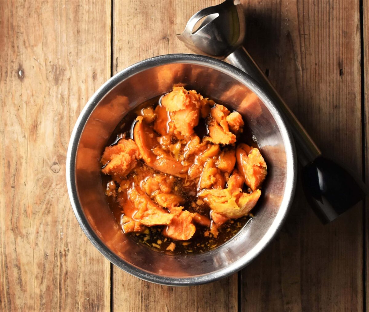 Mashed sweet potato and maple syrup in metal bowl with stick blender end piece in background.