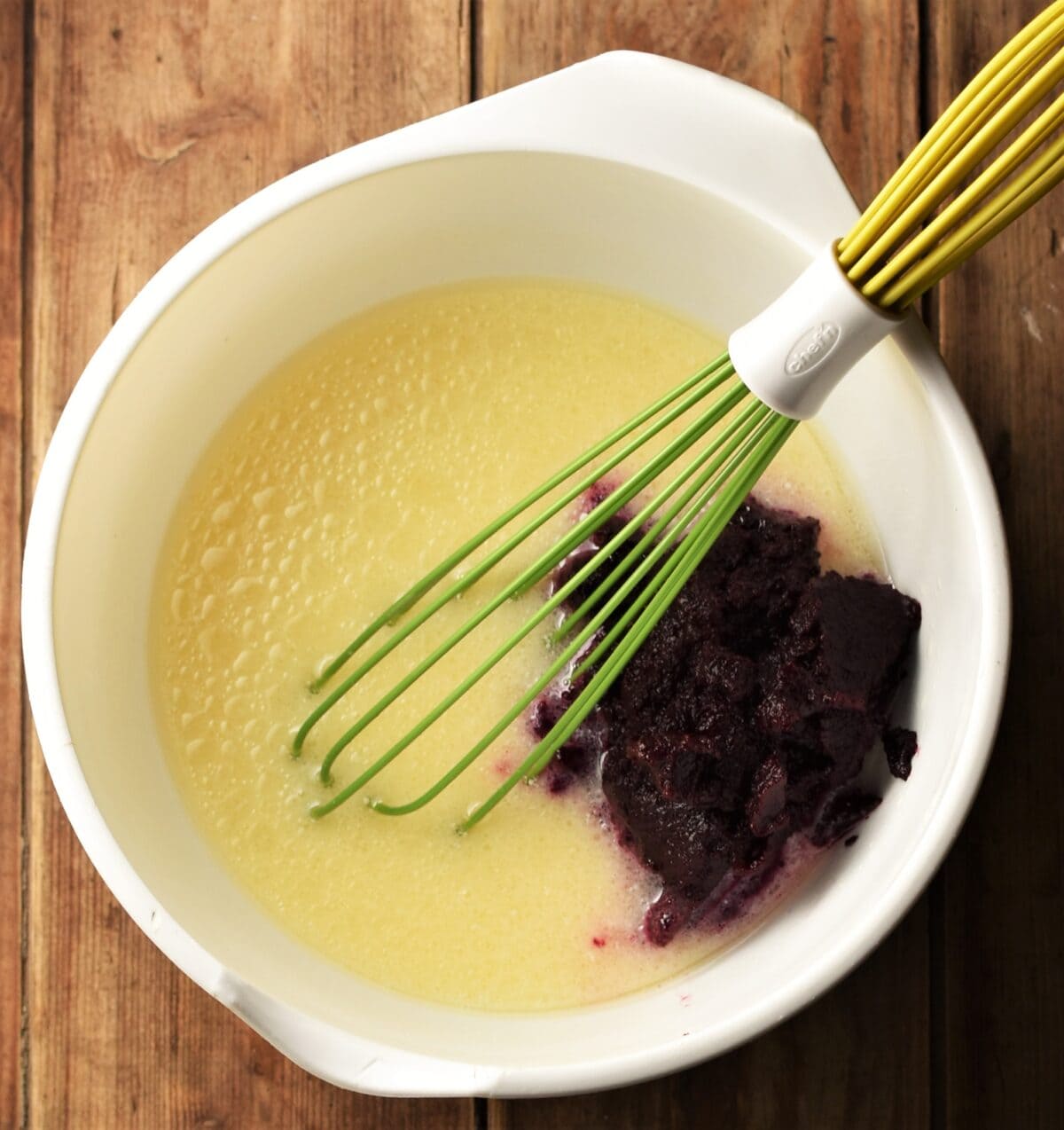 Egg mixture and beetroot puree in large white bowl with green whisk.
