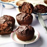 Side view of beetroot chocolate muffins on top of small white plate and inside ceramic muffin pan, with knife in background.