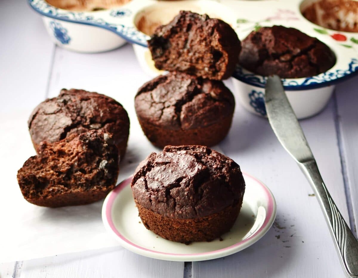 Side view of beetroot chocolate muffins on top of small white plate and inside ceramic muffin pan, with knife in background.