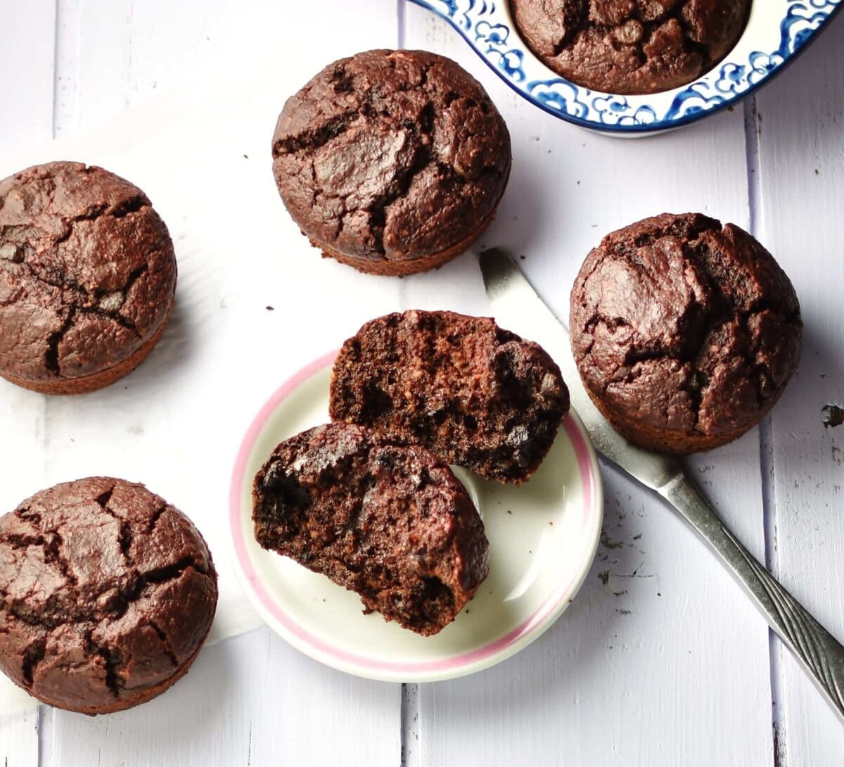 Top down view of chocolate muffins on top of small white plate, paper and inside ceramic muffin pan in background.