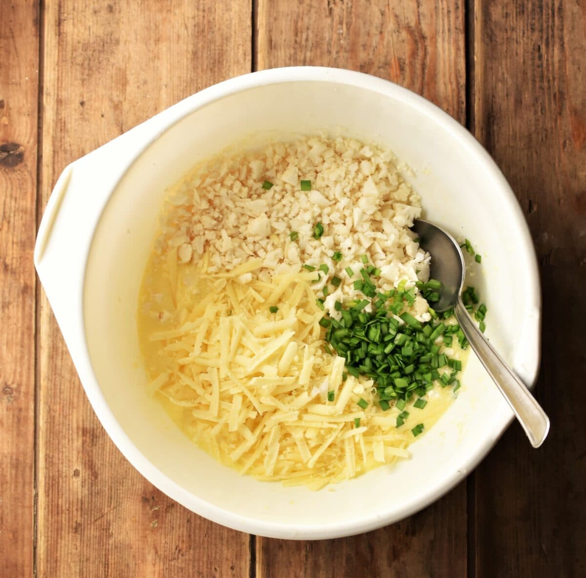 Grated cheese and cauliflower rice with chopped chives in large white bowl with spoon.