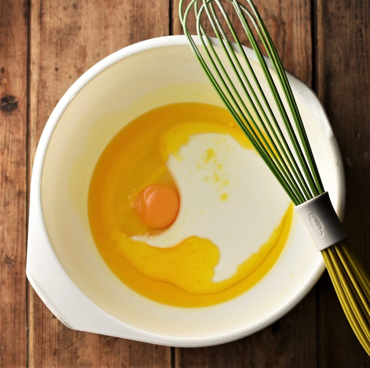 Egg with oil and milk in large white bowl with green whisk.