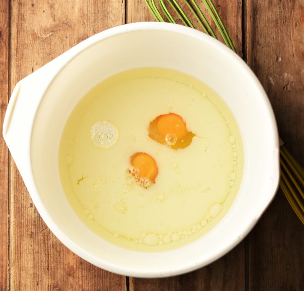 Milk, 2 eggs and oil in large white bowl with green whisk in background.