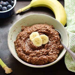 Quinoa chocolate porridge with slices of banana and spoon in light green bowl, with green cloth, banana and blueberries in small dish in background.