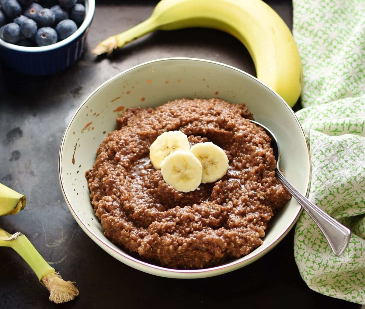 Quinoa chocolate breakfast porridge with slices of banana and spoon in light green bowl, with green cloth, banana and blueberries in small dish in background.