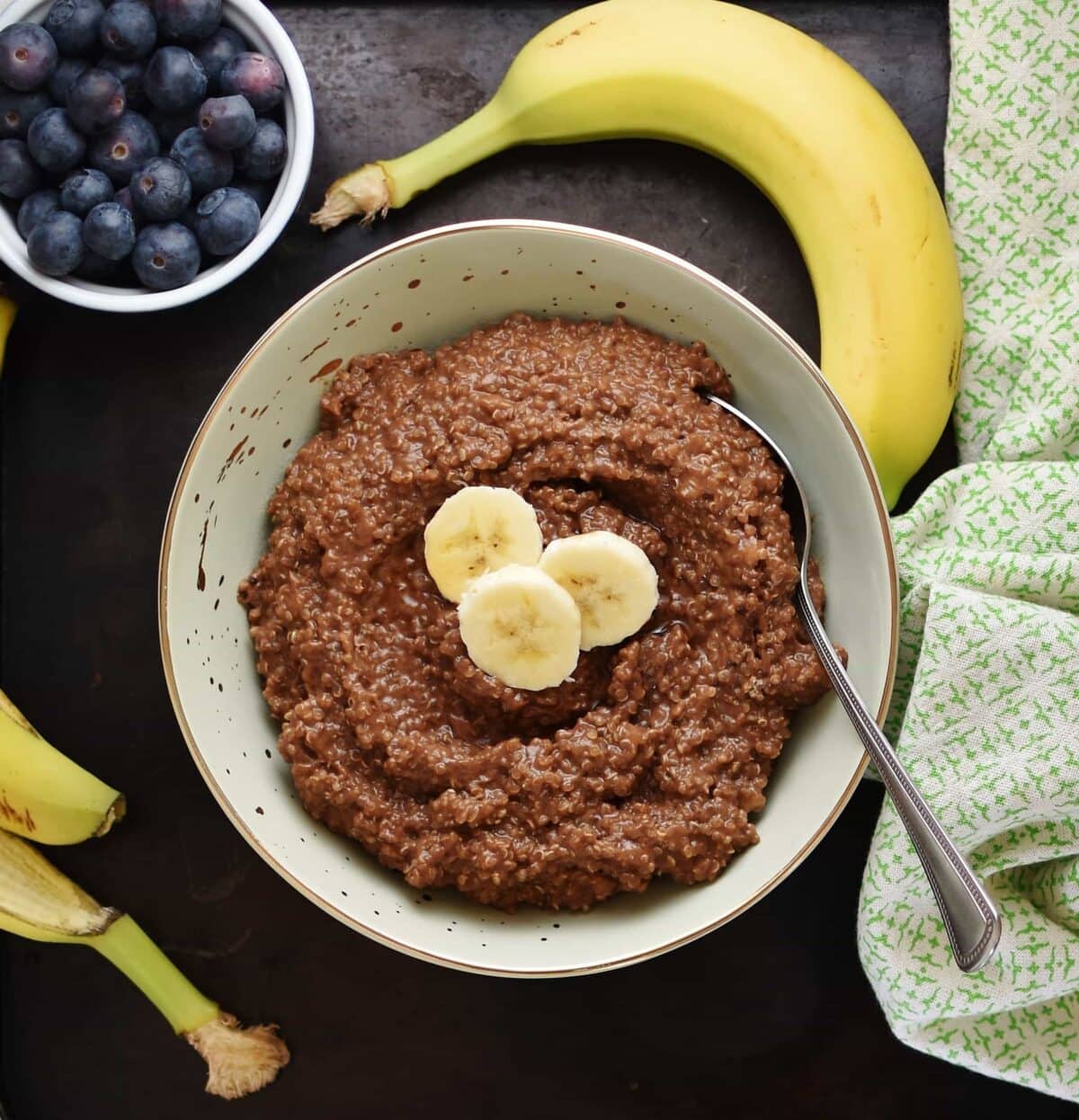 Top down view of quinoa breakfast porridge with slices of banana and spoon in light green bowl, with green cloth, banana and blueberries in round dish in background.
