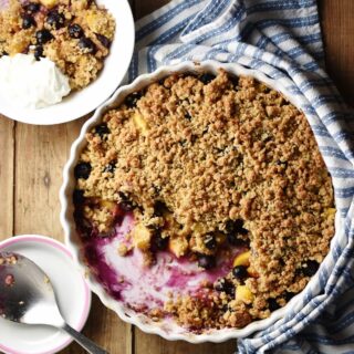 Top down view of peach and blueberry crumble inside white round ceramic dish wrapped in blue-and-white stripy cloth, with crumble, spoon and yogurt in white bowl and large spoon on top of small sauce in background.