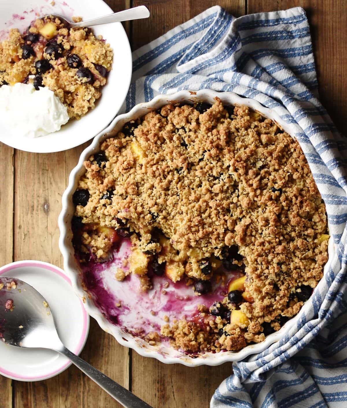 Top down view of peach and blueberry crumble inside white round ceramic dish wrapped in blue-and-white stripy cloth, with crumble, spoon and yogurt in white bowl and large spoon on top of small sauce in background.