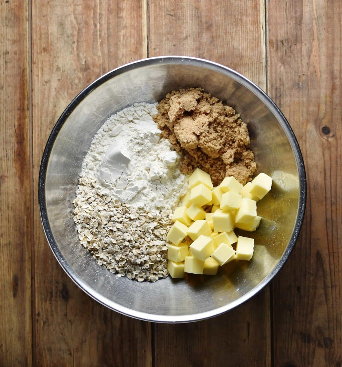 Top down view of oats, flour, sugar and cubed butter in metal bowl.