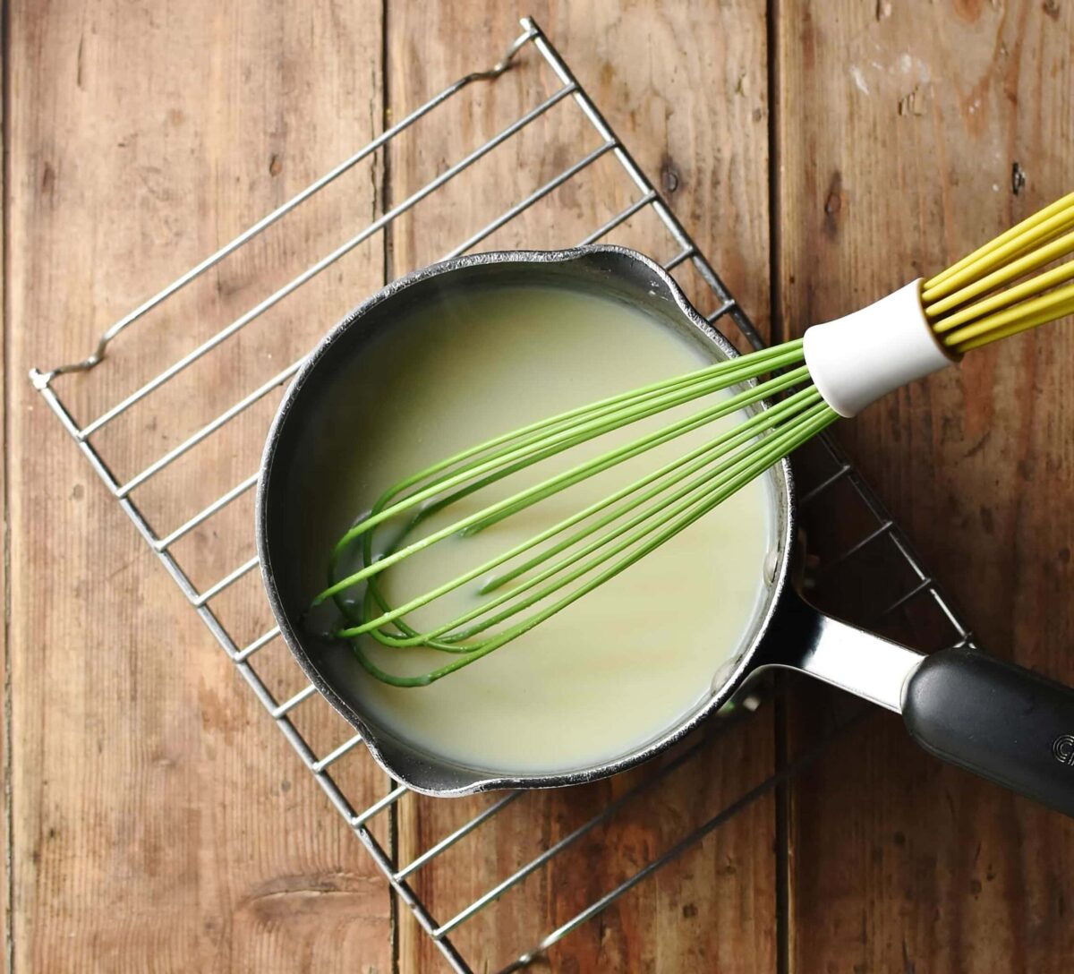 White sauce with green whisk in saucepan on top of rack.