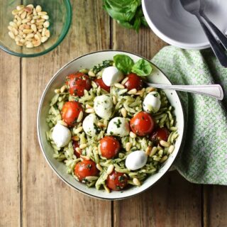 Top down view of orzo salad with pesto, cherry tomatoes, mini mozzarellas and spoon in white bowl wrapped in green cloth, with white bowl in top right and pine nuts in small dish in top left corner.