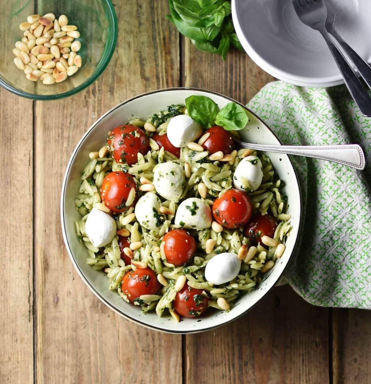 Top down view of orzo salad with pesto, cherry tomatoes, mini mozzarellas and spoon in white bowl wrapped in green cloth, with white bowl in top right and pine nuts in small dish in top left corner.
