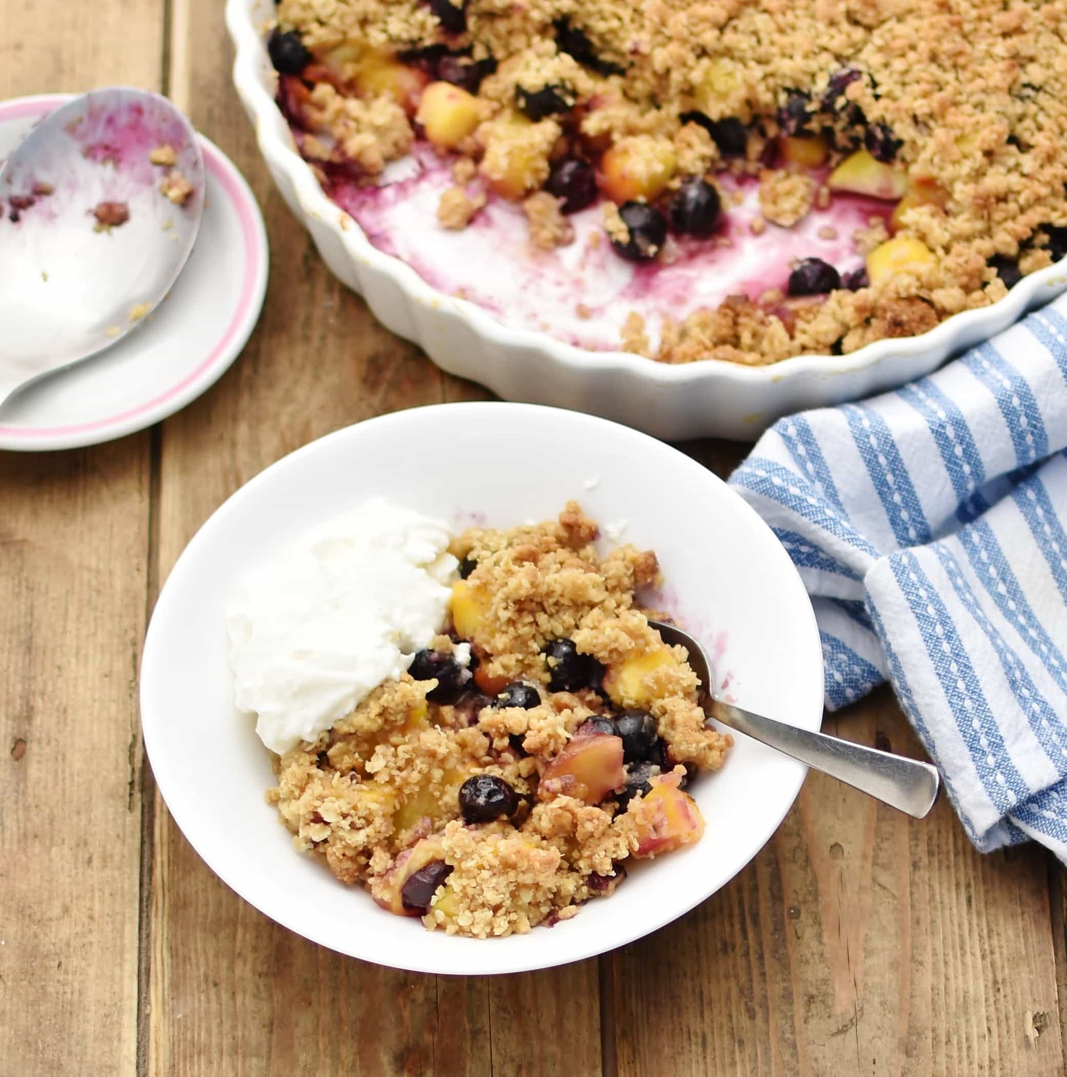 Blueberry peach crumble with yogurt and spoon inside white bowl, with partial view of white casserole dish containing crumble, wrapped in blue-and-white stripy cloth, and large spoon on top of saucer in background.