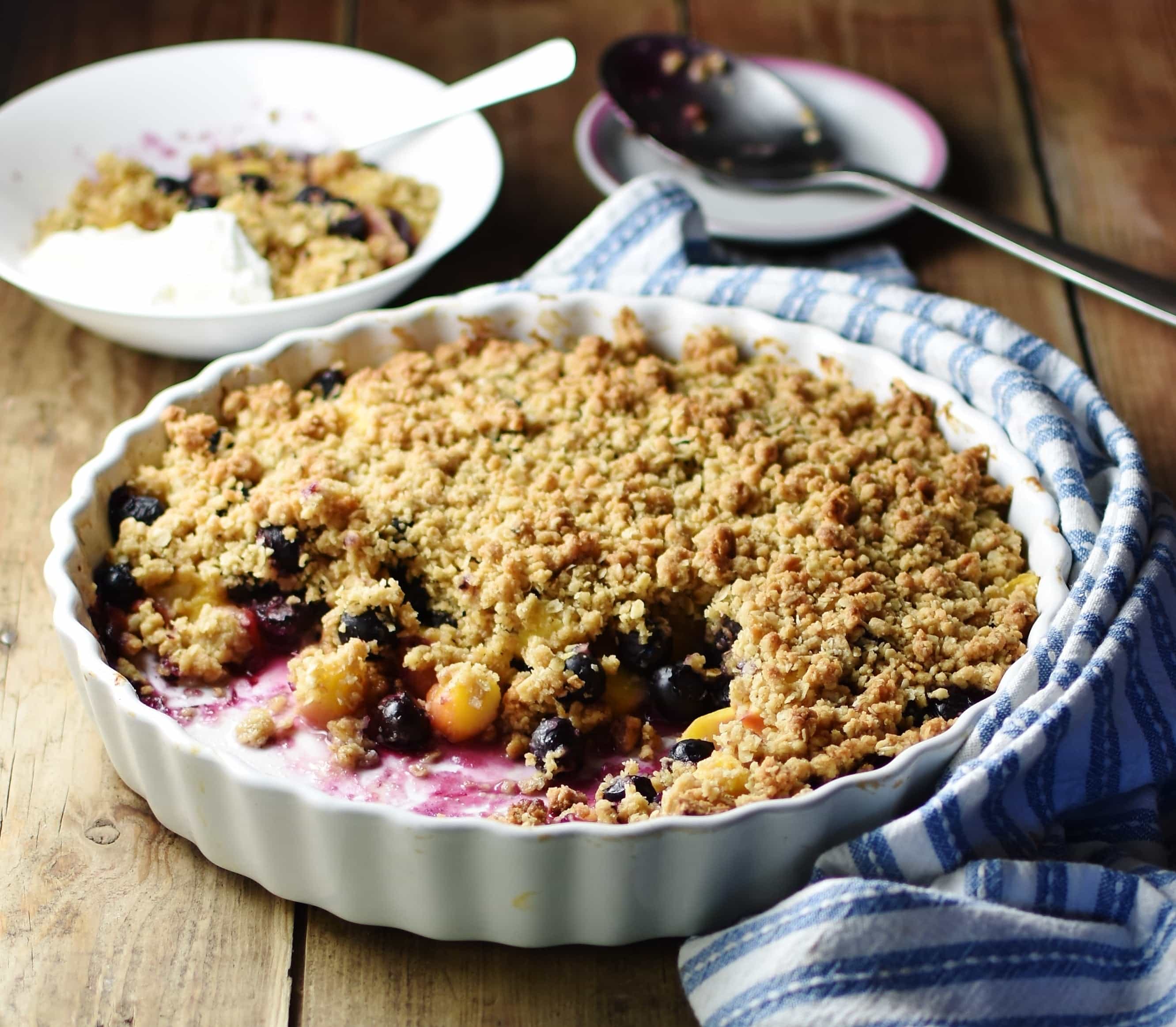 Side view of blueberry peach crisp in large white ceramic round dish wrapped in blue-and-white stripy cloth, with large spoon on top of small saucer, crumble in white bowl with spoon and yogurt in background.