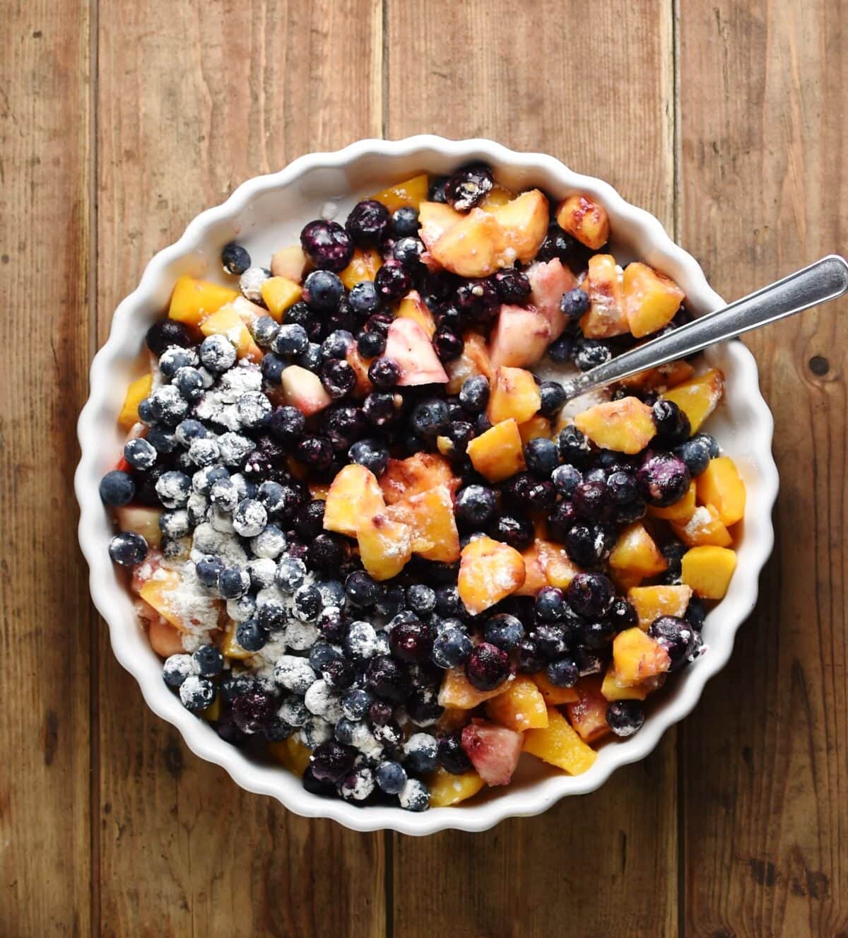 Top down view of cubed peaches and blueberries with flour and spoon in white pie dish.