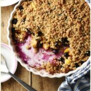 Top down view of peach and blueberry crumble inside white round ceramic dish wrapped in blue-and-white stripy cloth, with crumble, spoon and yogurt in white bowl and large spoon on top of small sauce in background.
