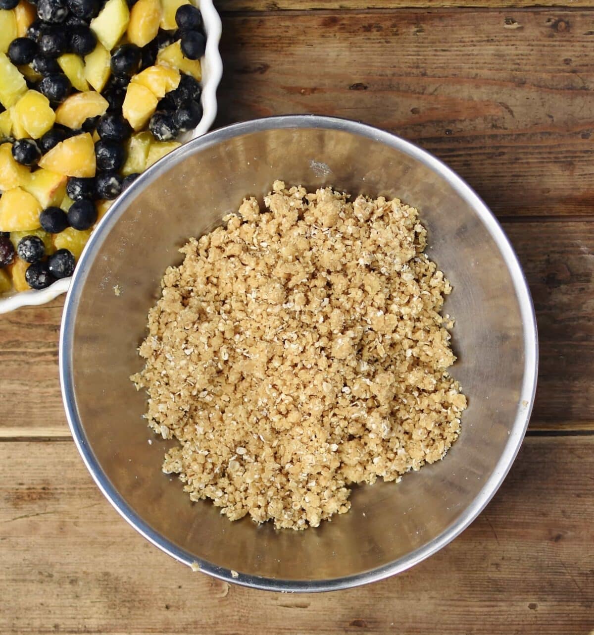 Top down view of crumble topping mixture inside metal bowl, with partial view of white casserole dish containing fruit in top left corner.