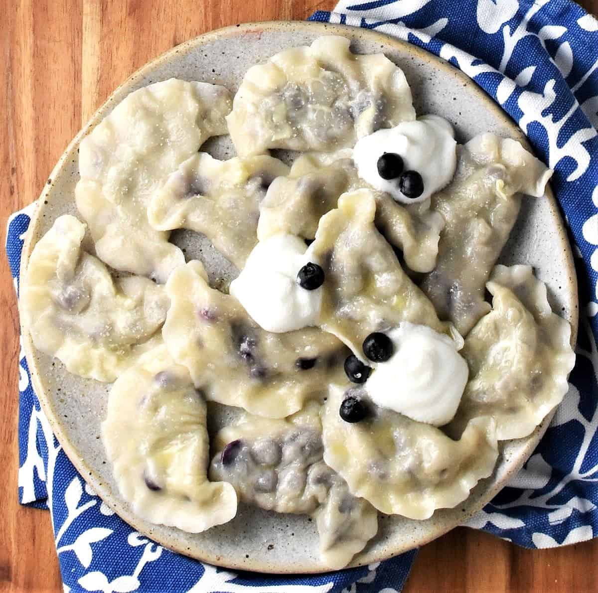 Top down view of Polish pierogi with blueberries and sour cream on plate with blue cloth in background.