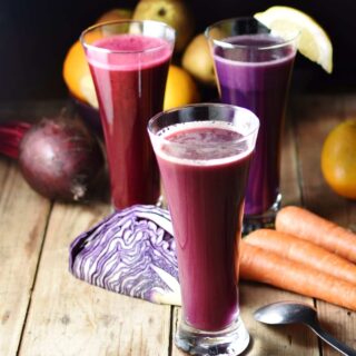 Side view of cabbage juice in 3 separate tall glasses with red cabbage, carrots, beet, and other raw vegetables and fruits in background on top of wooden board with spoon to the right.