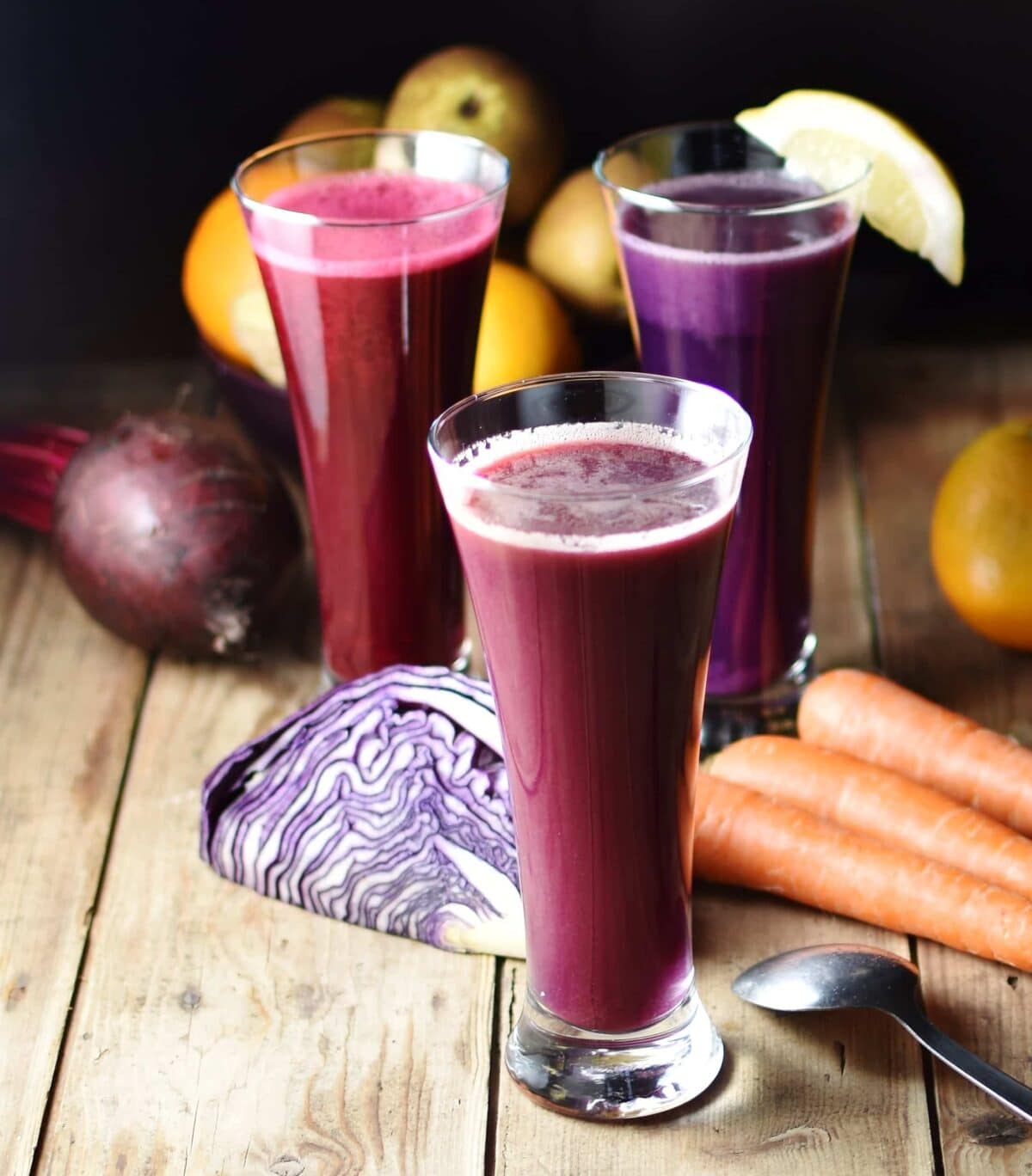Side view of cabbage juice in 3 separate tall glasses with red cabbage, carrots, beet, and other raw vegetables and fruits in background on top of wooden board with spoon to the right.