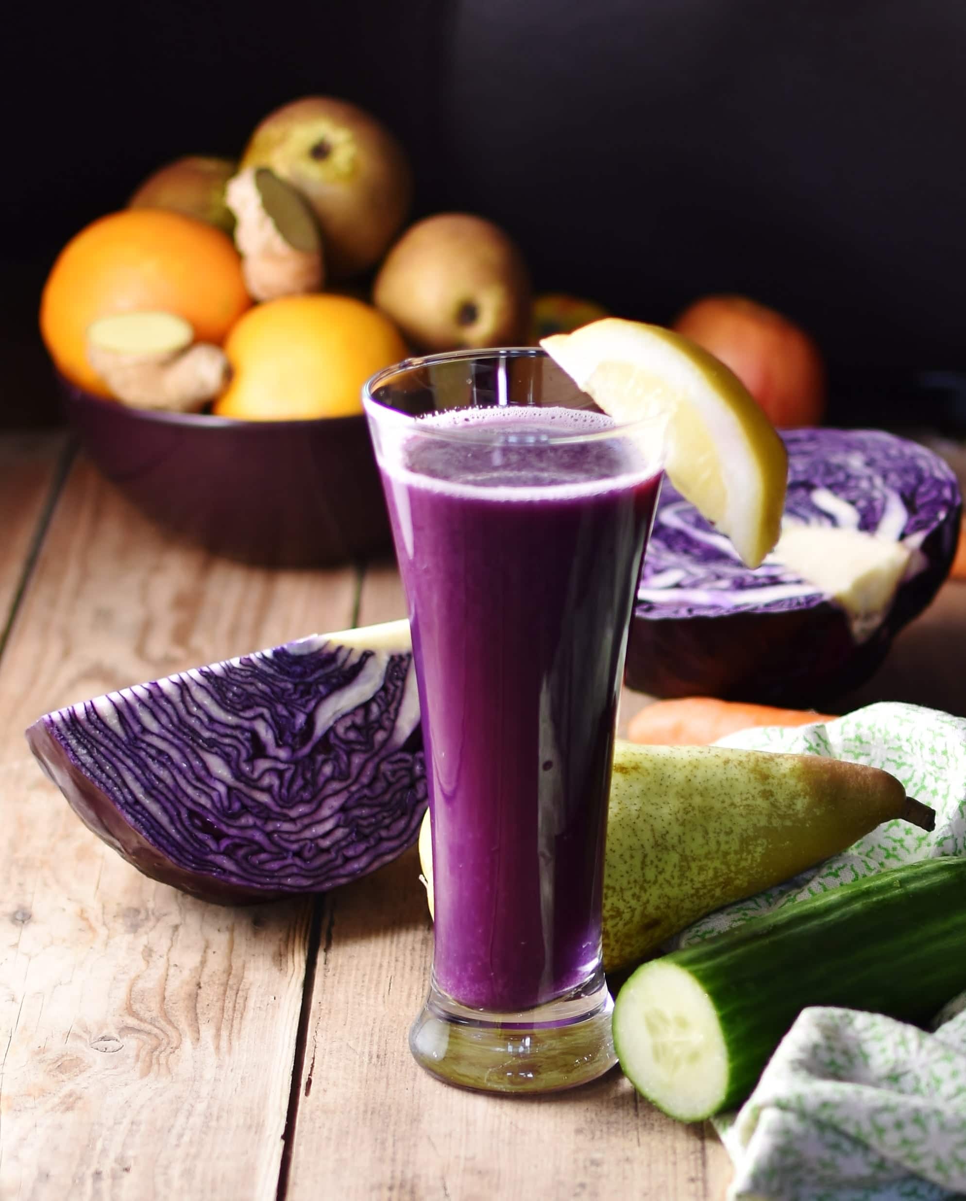 Side view of red cabbage juice inside tall glass with lemon wedge, with red cabbage, pear and cucumber on top of green cloth and more vegetables and fruits in background.